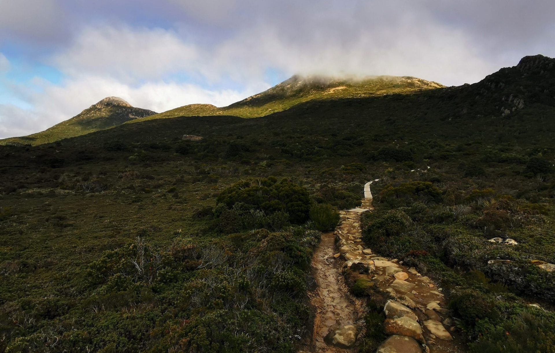 Lanscape photo of a mountain behind low-hanging clouds. A rocky path in the foreground resembles a lizard's tail.