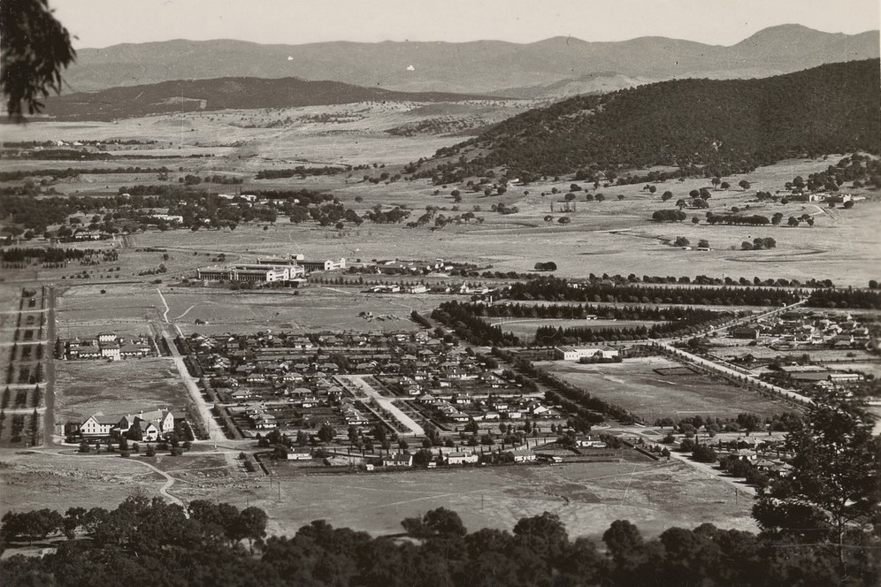 A greyscale photograph of the view of Canberra from Mount Ainslie in the 1930s. It shows a handful of tightly gridded suburbs surrounded by bushland. There are hills and mountains in the background.